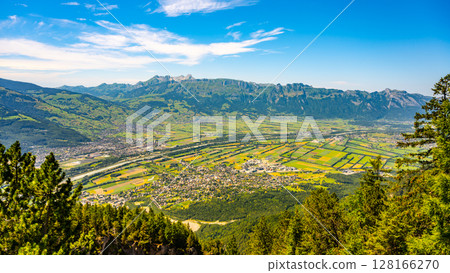 A stunning vista from Gaflei lookout tower showcases the picturesque town of Schaan, surrounded by lush greenery and mountain ranges under a clear blue sky. A stunning vista from Gaflei lookout tower showcases the picturesque town of Schaan, surrounded by lush greenery and mountain ranges under a clear blue sky. 128166270