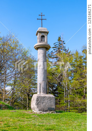 A tall stone column with a cross at the top rises from a lush green field surrounded by trees. The sky is clear and bright, emphasizing the peaceful yet haunting presence of the structure. 128166271
