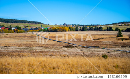 A vast autumn landscape unfolds at Bozi Dar peat bog, featuring golden grasses and a distant view of colorful trees and settlements under a clear blue sky in the Ore Mountains. A vast autumn landscape unfolds at Bozi Dar peat bog, featuring golden grasses and a distant view of colorful trees and settlements under a clear blue sky in the Ore Mountains. 128166299