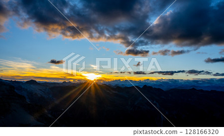 A stunning sunset unfolds over towering mountains, illuminating the sky with vibrant hues of orange and gold. Clouds drift lazily as the sun dips below the horizon, creating a captivating sight. A stunning sunset unfolds over towering mountains, illuminating the sky with vibrant hues of orange and gold. Clouds drift lazily as the sun dips below the horizon, creating a captivating sight. 128166320
