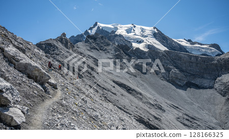 A group of hikers trek along a rocky trail with the snow-capped peak of Ortles mountain towering in the background under a clear blue sky. 128166325