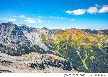 The winding roads of Stelvio Pass cut through the Italian Alps, showcasing a blend of rugged peaks and summer greenery against a clear blue sky. 128166327