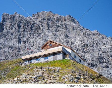 A traditional alpine hut, the Tabaretta Hut, is nestled on a lush green slope beneath the imposing Ortles mountain under a clear blue sky. A traditional alpine hut, the Tabaretta Hut, is nestled on a lush green slope beneath the imposing Ortles mountain under a clear blue sky. 128166330