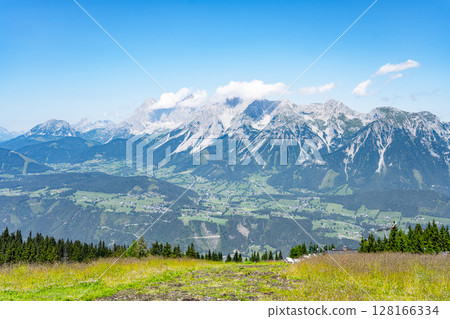Stunning panorama of the Dachstein mountain range viewed from Planai, Austria. Lush greenery contrasts with majestic peaks under a clear blue sky, creating a serene summer landscape. Stunning panorama of the Dachstein mountain range viewed from Planai, Austria. Lush greenery contrasts with majestic peaks under a clear blue sky, creating a serene summer landscape. 128166334