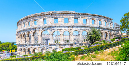 The Roman amphitheatre in Pula showcases ancient architecture amidst lush greenery. Visitors admire this historical site under a clear blue sky, reflecting on its rich past. The Roman amphitheatre in Pula showcases ancient architecture amidst lush greenery. Visitors admire this historical site under a clear blue sky, reflecting on its rich past. 128166347