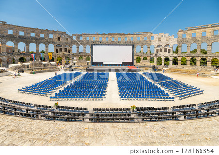 In the historic Roman amphitheatre of Pula, rows of blue chairs set the stage for an outdoor film screening. The ancient architecture creates a unique backdrop under a clear blue sky. In the historic Roman amphitheatre of Pula, rows of blue chairs set the stage for an outdoor film screening. The ancient architecture creates a unique backdrop under a clear blue sky. 128166354