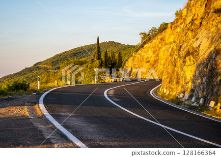 The winding coastal road stretches gracefully along the hillside, illuminated by the warm glow of the setting sun, surrounded by lush greenery and rocky formations. 128166364
