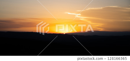The warm hues of a summer sunset blend beautifully as the sun dips below the horizon. Silhouettes of hills create a serene backdrop, highlighting the tranquility of the evening sky. 128166365