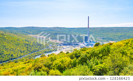 Plomin Power Station is set against a vibrant green backdrop on the Istria Peninsula in Croatia. The smokestack rises prominently, surrounded by hills and vegetation under a clear sky. 128166423