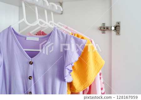 colorful children's clothes, including a lilac dress with ruffled sleeves, hangs on white hangers inside closet 128167059