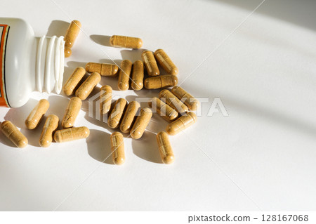 A white pill bottle lays on its side with brown capsules spilling out onto a clean white surface 128167068