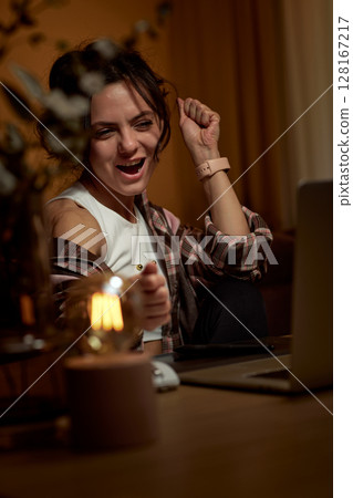 happy excited graphic designer woman with open mouth raised fist up working using digital graphic tablet at home in the evening 128167217