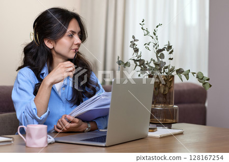 portrait of woman in blue shirt holds folders uses laptop in home office portrait of woman in blue shirt holds folders uses laptop in home office 128167254
