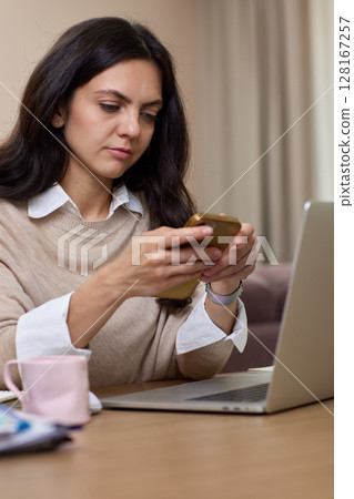 portrait of woman holds smart phone uses laptop in home office portrait of woman holds smart phone uses laptop in home office 128167257