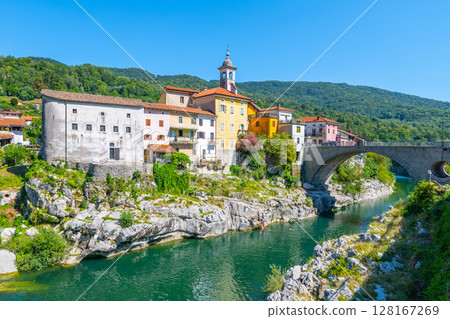 Kanal bridge gracefully stretches over the clear waters of the Soca River, surrounded by charming houses nestled against a backdrop of lush green hills on a sunny day. 128167269
