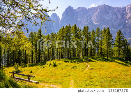 Explore the stunning landscape of Prisank Mountain in the Julian Alps, Slovenia, featuring a rock window, lush green meadows, towering trees, and scenic pathways under a bright sky. 128167278