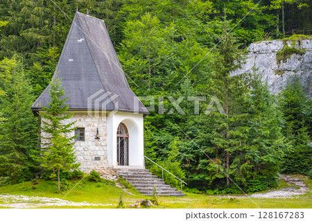 A quaint chapel stands amid lush greenery in Triglav National Park, Slovenia. Surrounded by tall trees, it offers a peaceful retreat and stunning natural scenery. 128167283