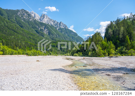 A serene view of a parched riverbed with patches of water, surrounded by lush forests and the towering peaks of the Julian Alps in Triglav National Park. Jasna near Kranjska Gora, Slovenia 128167284