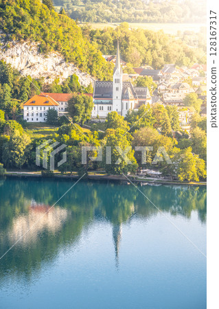 St. Martin's Church stands gracefully on a hillside, overlooking the tranquil waters of Lake Bled. Trees and mountains create a serene backdrop during golden hour. 128167317