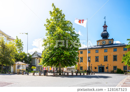Vaxholm Island Town Hall stands prominently with its unique architecture, surrounded by trees and flags. The sun shines brightly, creating a picturesque view of the town square. 128167323