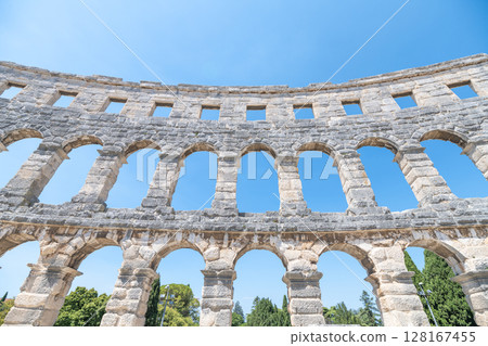 Visitors admire the ancient Roman amphitheatre in Pula, Istria Peninsula, showcasing its impressive arches and stonework under the clear blue sky, a testament to historical architecture. 128167455