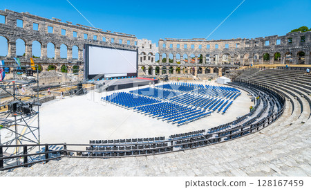 The Roman amphitheatre in Pula, Croatia, hosts an outdoor event setup with rows of seating and a large screen. Sunlight bathes the ancient stone structure, highlighting its history and beauty. 128167459