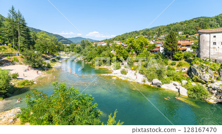 A tranquil stretch of Soca River meanders through Kanal, Slovenia, with green hills rising on either side. Nearby, people enjoy the clear water and warm sunshine, creating a lively atmosphere. 128167470