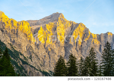 Golden sunlight illuminates Triglav Mountain in the Julian Alps. The majestic peak rises sharply against the clear blue sky, surrounded by serene forested foothills. Golden sunlight illuminates Triglav Mountain in the Julian Alps. The majestic peak rises sharply against the clear blue sky, surrounded by serene forested foothills. 128167486