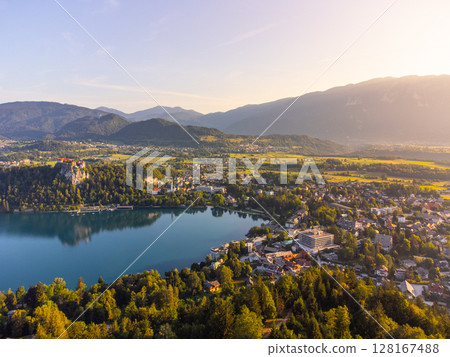 Bled Lake sparkles under the golden sunlight, framed by green forests and mountains. The charming town of Bled is visible, with its historic castle perched on a cliff. 128167488