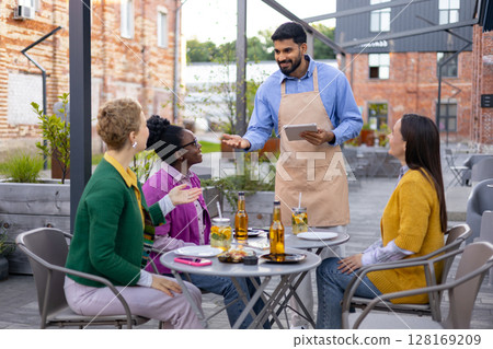 A waiter takes an order from three women seated at an outdoor restaurant table. They are enjoying drinks and conversation. A waiter takes an order from three women seated at an outdoor restaurant table. They are enjoying drinks and conversation. 128169209