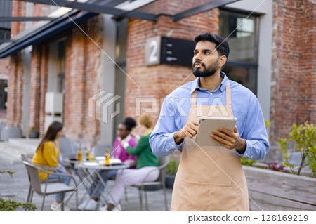 A Hindu male waiter holds a tablet, looking away from the camera at an outdoor restaurant. People sit at a table in the background. 128169219