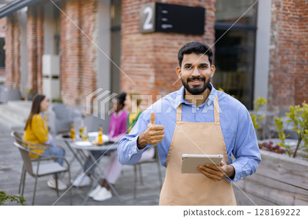 A Hindu male waiter with a beard smiles and holds a tablet, giving a thumbs-up at an outdoor restaurant. 128169222