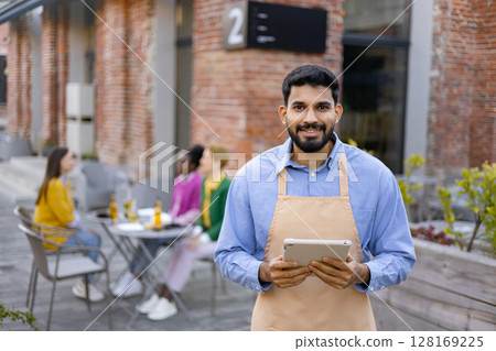 A smiling Hindu male waiter in an apron holds a tablet, standing outside a restaurant. 128169225