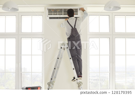 Service worker wearing overalls, standing on ladder, repairing air conditioner Service worker wearing overalls, standing on ladder, repairing air conditioner 128170395