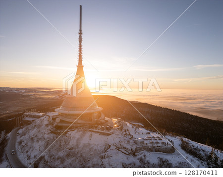 At sunrise, the snow-covered Jested Mountain showcases its unique architecture against a backdrop of golden light. The landscape is tranquil, with a sense of calm enveloping the peak. 128170411