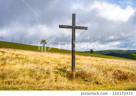 A wooden cross rises prominently in a mountain meadow near Haj, Czechia. Rolling hills and a solitary tree frame this serene landscape under a partly cloudy sky. 128170433