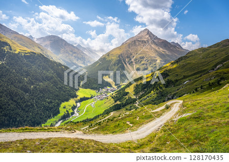 Beautiful landscape captures Vent village nestled in the Otztal Alps. Lush green valleys extend below, surrounded by majestic mountains under a clear blue sky with soft clouds. 128170435