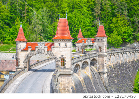A view of the Les Kralovstvi Dam in Czechia, showcasing the intricate archway and stonework of the structure. The dam stands tall and imposing, surrounded by lush greenery. A view of the Les Kralovstvi Dam in Czechia, showcasing the intricate archway and stonework of the structure. The dam stands tall and imposing, surrounded by lush greenery. 128170456