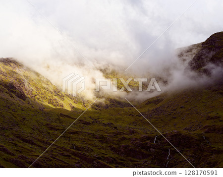 Misty clouds drift through the valley of Carrauntoohill. Low-hanging clouds obscure the mountains peak, adding to the dramatic atmosphere of the Irish landscape. 128170591