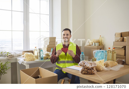 Smiling young volunteer holding red heart in hands sitting at charity center packing donations 128171070