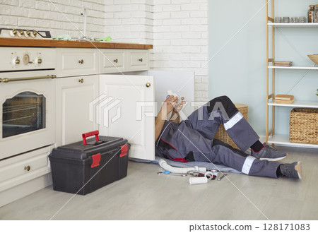Male plumber in work uniform lying on floor, fixing kitchen sink pipes, providing plumbing service. Male plumber in work uniform lying on floor, fixing kitchen sink pipes, providing plumbing service. 128171083