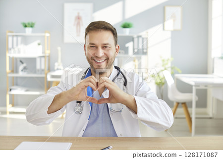 Smiling male doctor in white coat sitting at desk, making heart shape with hands in medical office. 128171087