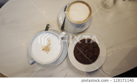 Two cups of cappuccino with milk foam served with a chocolate cookie on a white plate. This image represents a cozy coffee break and sweet indulgence concept 128171140