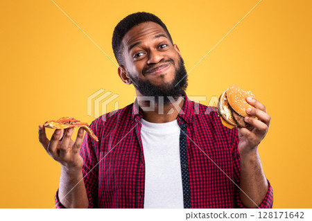 Smiling African Man Holding Burger And Slice Of Pizza Posing Standing In Studio Over Yellow Background. Junk Food Eater's Portrait. Unhealthy Fastfood And Male Nutrition, Cheat Meal And Overeating 128171642