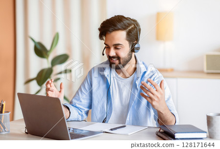 Web Conference. Friendly Arabic Freelancer Guy In Headset Making Video Call On Laptop, Sitting At Desk In Home Office, Talking And Gesturing At Camera, Enjoying Online Communication, Copy Space 128171644