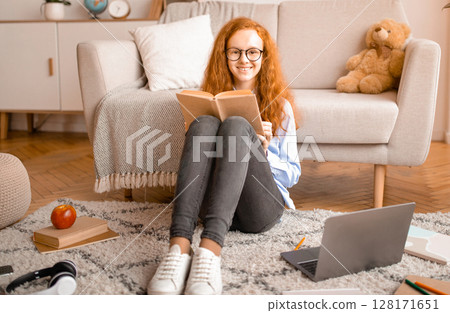 Education Concept. Full length portrait of cute smiling red-haired girl wearing glasses, sitting on the floor carpet with laptop, reading paper book, looking at camera. Teen resting at home 128171651