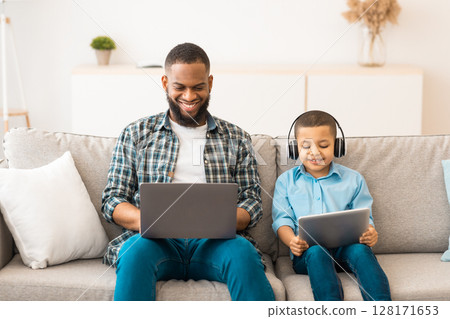 Black Father And Little Boy Using Computers Together Browsing Internet On Laptop And Digital Tablet Wearing Wireless Headphones Sitting On Sofa At Home. Family Gadgets Concept 128171653