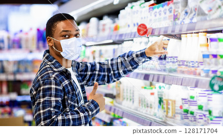 Millennial black guy in facial mask taking bottle of milk from supermarket shelf, gesturing thumb up, recommending good grocery store, shopping at mall during coronavirus lockdown Millennial black guy in facial mask taking bottle of milk from supermarket shelf, gesturing thumb up, recommending good grocery store, shopping at mall during coronavirus lockdown 128171704