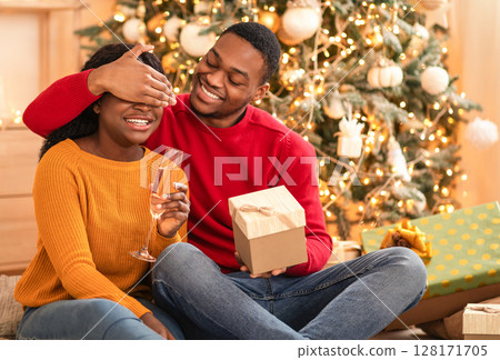 Presents time in living room. Smiling young african american husband closes eyes to woman with glass of champagne and gives box in home interior with Christmas tree with glowing garlands, copy space Presents time in living room. Smiling young african american husband closes eyes to woman with glass of champagne and gives box in home interior with Christmas tree with glowing garlands, copy space 128171705