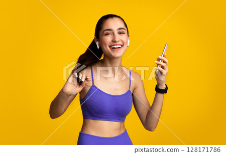 Music And Sport. Portrait Of Excited Young Fit Woman In Sportswear And Wireless Earbuds Dancing Isolated Over Orange Studio Background And Enjoying Favorite Song, Holding Cellphone Music And Sport. Portrait Of Excited Young Fit Woman In Sportswear And Wireless Earbuds Dancing Isolated Over Orange Studio Background And Enjoying Favorite Song, Holding Cellphone 128171786
